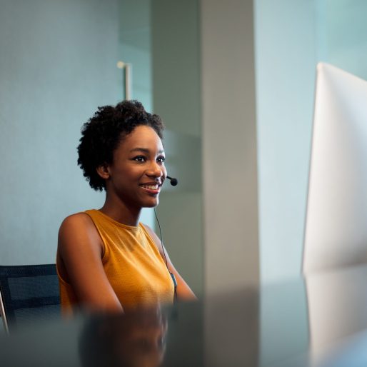 A beautiful young female receptionist sitting in front of a computer, wearing a headset and smiling.